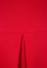 Červené šaty s texturovanou tkaninou, s vertikálním plisováním uprostřed, vytvářející čistou a strukturovanou siluetu.