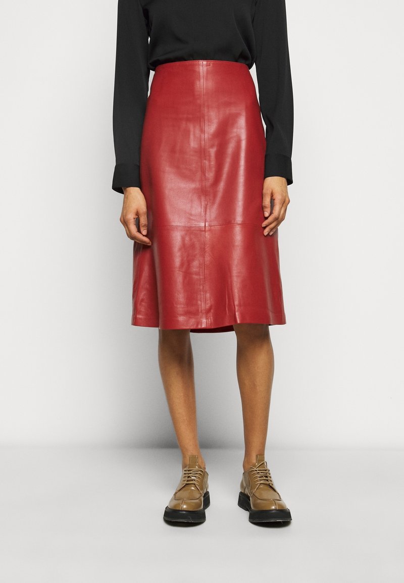 Femme portant une jupe en cuir rouge longueur genou, un haut noir à manches longues et des chaussures à lacets couleur beige, debout devant un fond uni.