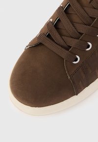 Κοντινό πλάνο ενός καφέ αθλητικού παπουτσιού με ταιριαστά κορδόνια, μεταλλικά ματιά και μπεζ καουτσούκ σόλα σε λευκό φόντο.