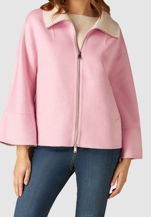 Femme portant une veste zippée rose clair avec de larges manches, un col crème, un top beige en dessous et un jean bleu sur un fond uni.
