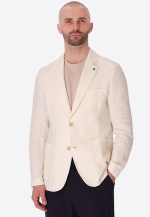 Uomo con barba rifilata che indossa un blazer beige sopra una camicia color sabbia chiaro, in piedi con una mano in tasca davanti a uno sfondo uniforme.