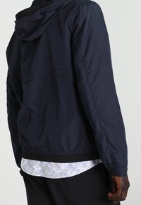 Navy hooded jacket with a smooth, lightweight fabric. Features elastic cuffs and hem. Worn over a patterned shirt, with a relaxed fit.