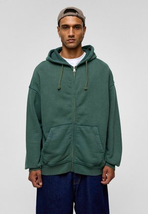 Groene rits-trui in een ontspannen pasvorm, met een trekkoordcapuchon, twee voorzakken en geribde manchetten. Draag het over een witte t-shirt.