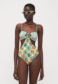 Maillot de bain une pièce multicolore avec un design ajusté, présentant un haut à carreaux verts, des motifs floraux et une fermeture à lien ajustable sur le devant.