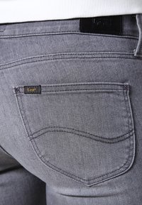 Jeans de mezclilla gris con un bolsillo trasero que presenta una pequeña etiqueta "Lee" en negro y dorado, acentos de costura sutiles y una textura suave.