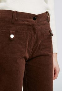 Pantalons en velours côtelé marron avec un tissu texturé, poches avant et détails en matériel argenté, dotés d'une taille haute et d'une coupe ajustée.