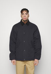 Marineblauwe, knoopsluiting jas met een zwarte kraag, twee voorzakken en een gladde textuur. Draagt met lichte broek.