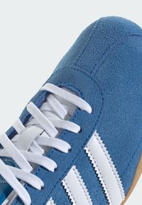 Baskets en nubuck bleu avec trois bandes blanches en cuir, tige texturée, lacets blancs et semelle en caoutchouc beige. Détails en zigzag remarquables sur les bandes.