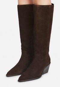 Alti stivali in suede marrone scuro con punte affusolate e tacchi medi inclinati, indossati da una persona su uno sfondo bianco uniforme.