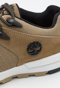 Baskets marron clair présentant un mélange de tissu texturé et de daim, avec des accents noirs et un logo circulaire, complétées par une semelle blanche.