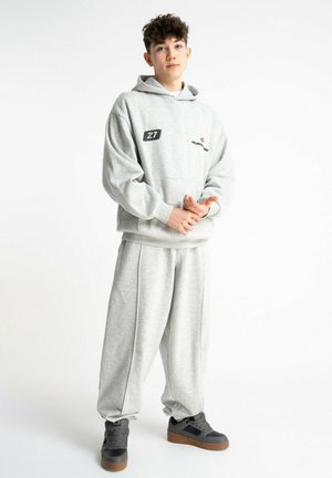 Młoda osoba stoi w jasnoszarej bluzie z kapturem i pasujących szarych spodniach dresowych oraz ciemnoszarych sneakersach, na tle jednolitej białej ściany.