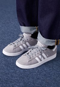 Sneakers Adidas Campus grigi con strisce bianche indossati con jeans blu scuro risvoltati su un pavimento blu in moquette.