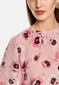 Blumenbluse aus zartem rosa Stoff mit großen, schwarz-gehörnten rosa Blumen und einem runden Ausschnitt, mit durchsichtigen langen Ärmeln.