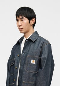 Τζιν μπουφάν σε σκούρο μπλε χρώμα με γιακά τύπου spread, χρυσά κουμπιά, δύο τσέπες στο στήθος και ένα μικρό λευκό λογότυπο στην αριστερή τσέπη.