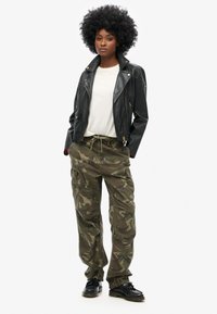 Zwarte leren motorjack over een witte t-shirt, gecombineerd met olijfgroene camouflage cargo shorts. Zwarte schoenen met een stevige zool.