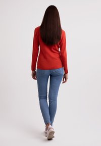 Röd långärmad tröja, blå tajta jeans och vita sneakers. Håret är långt och rakt, med en mjuk struktur. Vänder ryggen mot kameran.