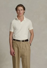 Baltā polo krekla ar īsām piedurknēm, apvienots ar brūņkrāsas šaha raksta biksēm un tumšu jostu. Modelis stāv ar rokām uz gurniem pret neitrālu fona.