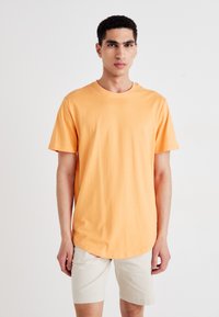 Trumpomis rankovėmis oranžinės spalvos medvilniniai marškinėliai su apvalia apačia, vilkimi su šviesiai smėlio spalvos šortais, pateikti ant gryno balto fono.