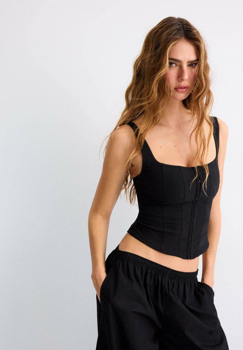 Jeune femme aux longs cheveux ondulés, vêtue d'un haut noir moulant sans manches et d'un pantalon noir ample, debout les mains dans les poches devant un fond uni.