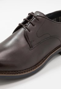 Chaussure de ville pour homme en cuir marron foncé à lacets, avec des coutures détaillées et une semelle superposée, sur fond blanc.