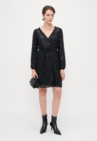 Vestido negro con lentejuelas, de mangas largas, escote en V y cintura ajustada, combinado con botas negras de tobillo y un bolso pequeño.