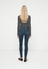 Jeans ajustados de mezclilla azul oscuro con cintura alta. La modelo lleva una blusa de rayas en blanco y negro de manga larga y unos zapatos planos marrones.
