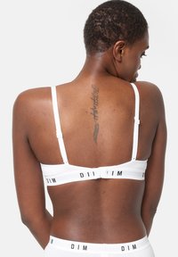 Una donna con i capelli corti che indossa un set di reggiseno e mutandine bianche con il marchio "DIM", mostrando un tatuaggio verticale sulla parte superiore della schiena.