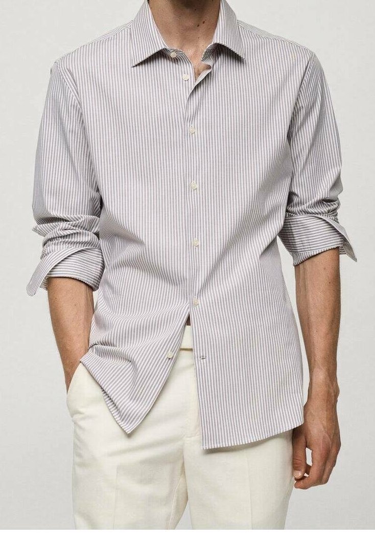 Homme portant une chemise à manches longues à rayures verticales grises claires et blanches avec les manches retroussées et un pantalon de couleur crème, debout avec une main dans la poche.