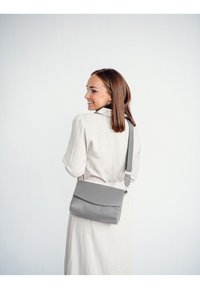 Šedá kožená crossbody taška s klopou a vyraženým logem, s nastavitelným popruhem a hladkou texturou.