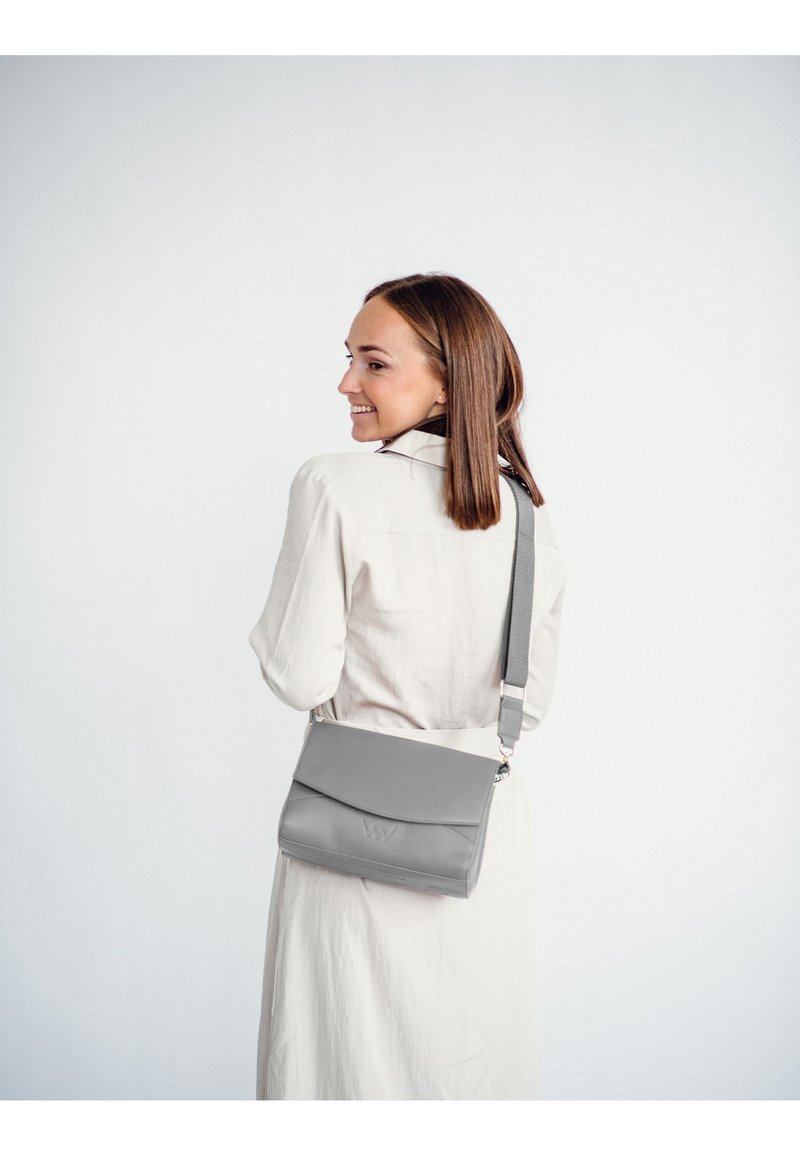 Šedá kožená crossbody taška s klopou a vyraženým logem, s nastavitelným popruhem a hladkou texturou.