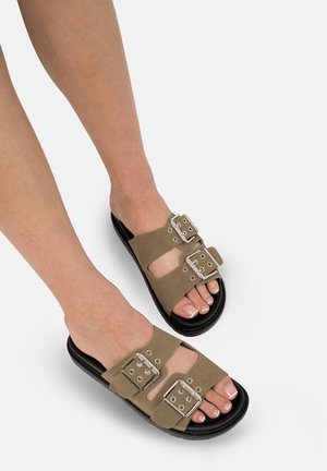 Sandali in suede marrone con doppie cinghie larghe, dotati di fibbie e dettagli metallici, poggiati su una suola in gomma nera. Design aperto davanti.