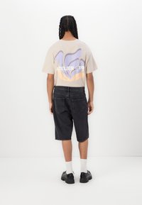 Beige t-shirt with purple and orange graphic on the back, paired with black denim shorts and chunky black shoes. White ankle socks visible.