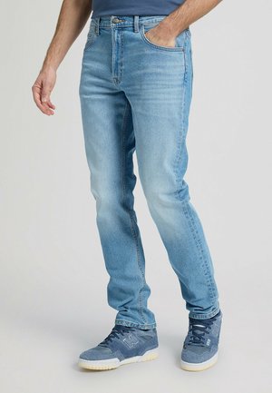 Mand iført lyseblå slim-fit jeans og mørkeblå sneakers, stående med den ene hånd i lommen mod en ensfarvet baggrund.