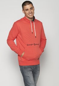 Červená mikina s kapucňou a textúrovaným dizajnom listov, sťahovacia kapucňa, predné vrecko a vyšívané logo. Spárovaná so sivej džínsami.