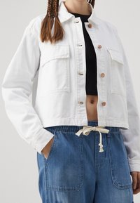 Vit kortärmad denimjacka med två framlommor, knappstängning och bronsbeslag, kombinerad med blå byxor med dragsko.