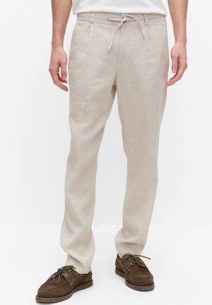 Man draagt beige linnen broek met trekkoord en bruine suède mocassin-schoenen, staand tegen een effen witte achtergrond.