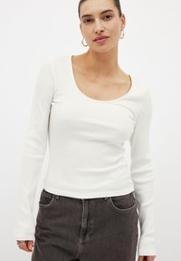 Top bianco a maniche lunghe con scollatura ampia a barca, caratterizzato da un design aderente e una texture liscia. Abbinato a jeans scuri.