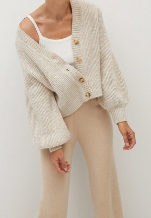 Kvinde iført en beige ribbet cardigan med træknapper over en hvid tanktop og beige tætsiddende bukser mod en ensfarvet lys baggrund.
