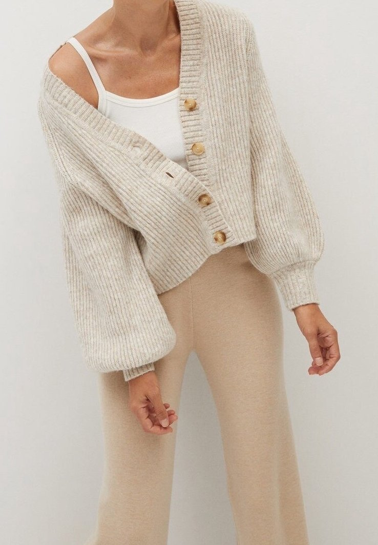 Femme portant un cardigan côtelé beige avec des boutons en bois sur un débardeur blanc et un pantalon beige ajusté, devant un fond clair uni.