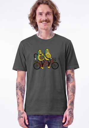 Homme avec tatouages et piercing au septum portant un t-shirt gris foncé avec des avocats dessinés faisant du tandem.