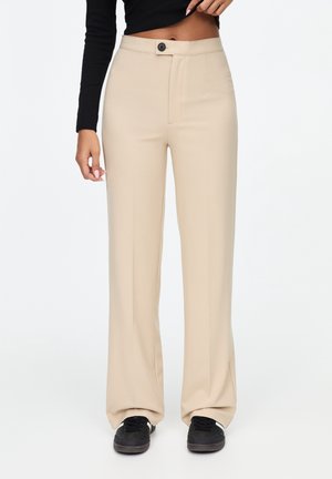 Frau trägt beige weite Hosen, die an der Taille mit Knöpfen versehen sind, schwarze Sneaker und ein kurzes schwarzes Langarmshirt.