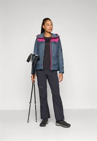 Žena u vanjskoj opremi drži planinarske štapove, nosi plavu i ružičastu jaknu, crnu majicu, tamne hlače i planinarske čizme, stoji na jednobojnoj pozadini.