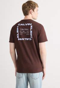 Camiseta de algodón marrón con un diseño gráfico en la parte posterior que incluye "Calvin Klein" y una dirección en texto blanco. Mangas cortas y cuello redondo.