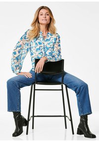 Hellblaues und beiges gemustertes Blusenshirt mit Ballonärmeln, getragen mit blauen Jeans und schwarzen Stiefeletten, sitzt auf einem schwarzen Stuhl.