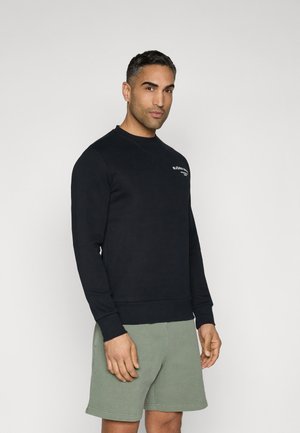 Mann trägt schwarzen langärmeligen Pullover und grüne Shorts, steht vor hellem, einfarbigem Hintergrund und blickt leicht nach rechts.