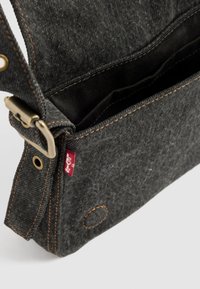 Sac en denim noir avec une poche intérieure, des coutures cuivrées, une boucle en métal et une petite étiquette logo sur le côté.