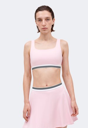 Žena oblečená v bledoružovom crop tope a zodpovedajúcej sukni s čierno-bielymi prúžkovanými elastickými pásmi, stojaca pred jednoduchým svetlým pozadím.