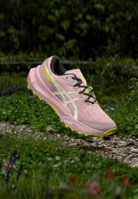 Zapatilla de correr color rosa claro con parte superior de malla texturizada, acolchado negro, acentos amarillos y cordones multicolores; diseñada para uso al aire libre.