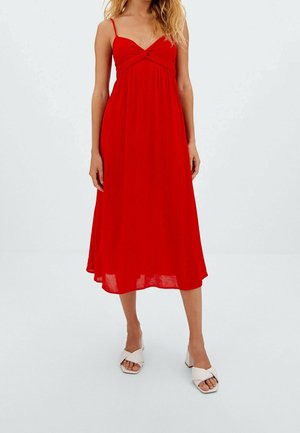 Femme portant une robe midi rouge sans manches à fines bretelles et des sandales blanches à talons, debout sur un fond blanc uni.