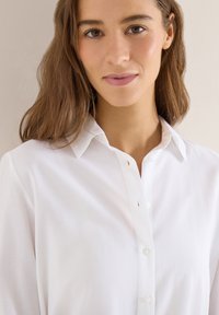 Jeune femme aux cheveux châtain clair portant une chemise blanche à boutons, souriant doucement contre un fond neutre.
